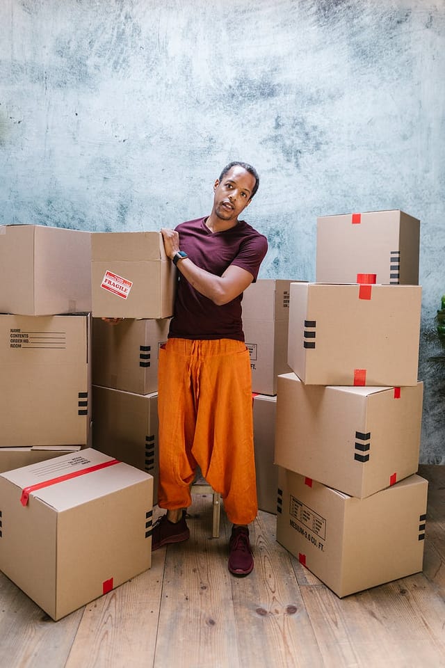 man in orange pants holding a package standing beside cardboard boxes- moving house planner